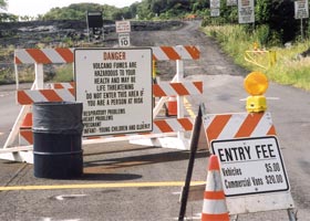 Two signs at beginning of lava viewing road: on sawhorse in foreground, 'Entry fee | Vehicles $5 | Commercial vans $20'; on barricade in background, 'Danger | Volcano fumes are | dangerous to your | health and may be | life threatening | Do not enter this area if | you are a person at risk | Respiratory problems | Heart problems | Pregnant | Infant-young children and elderly'
