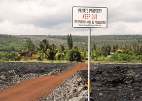 House in background at end of red dirt road through black lava field, behind 'No Trespassing' sign next to road